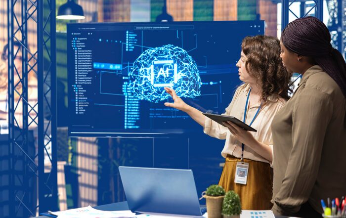 Two women on a tech team examine software with AI graphic on a screen behind them