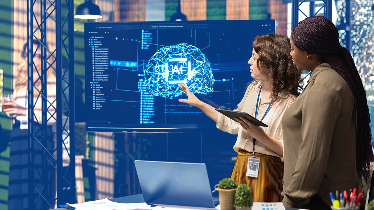 Two women on a tech team examine software with AI graphic on a screen behind them